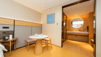 A modern hotel room with a round dining table, a small kitchenette, and a separate bathroom with a round mirror.