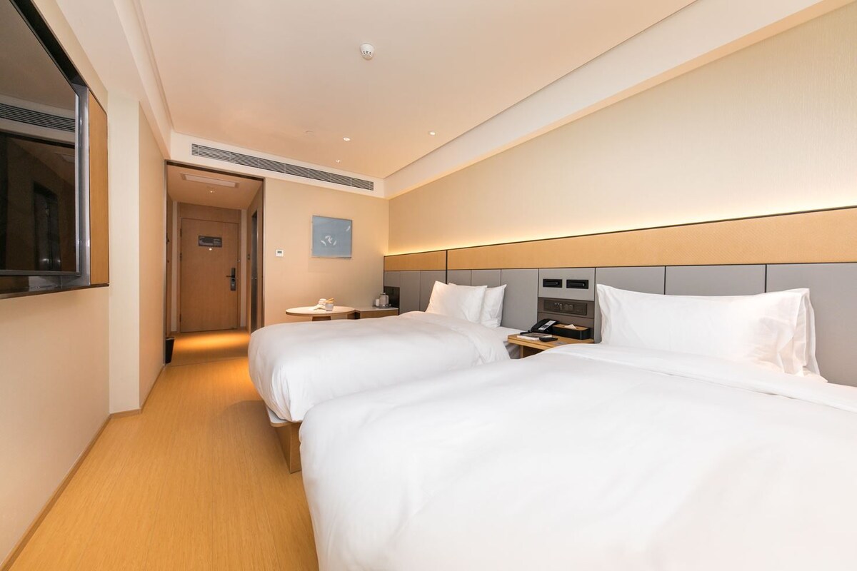 A hotel room with two beds, a bedside table with a phone and a lamp, a small table with a cup and a book, and a door leading to another room.