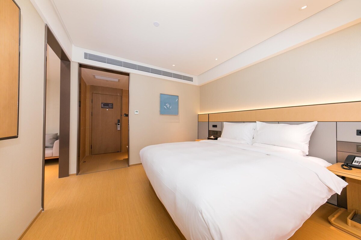A hotel room with a bed, a bedside table, a nightstand, a door, and a wall-mounted light fixture.