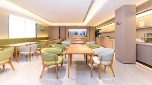 A modern dining area with a long table and chairs, a kitchen area with shelves, and a large window providing natural light. - Ji Hotel (Shanghai)