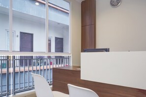 A reception area with a wooden reception desk, white chairs, and a large window offering a view of a building with numbered doors. - Super OYO 90115 Near Pasar Baru (Jakarta)