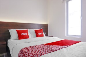 Una habitación de hotel con cama, dos almohadillas rojas con la palabra 'OYO' y una colcha blanca, además de una ventana que deja entrar luz natural.