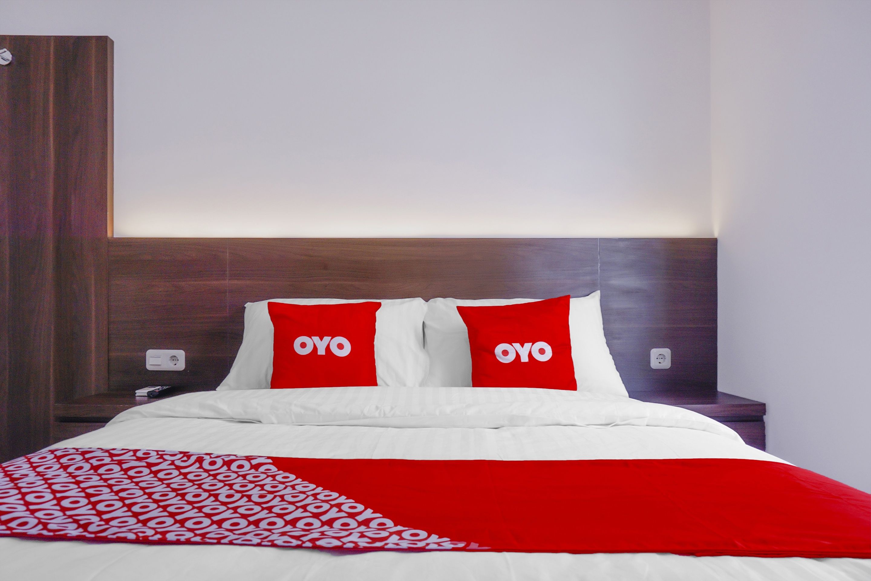 Une chambre d’hôtel avec un lit, deux oreillers rouges ornés du texte « OYO », une couverture blanche et rouge, et une tête de lit en bois.