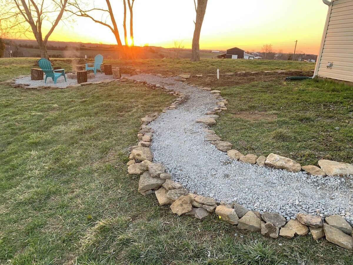 Un sentier de gravier bordé de rochers mène à un espace aménagé avec des chaises et un foyer, le tout se détachant sur le fond d’un coucher de soleil.