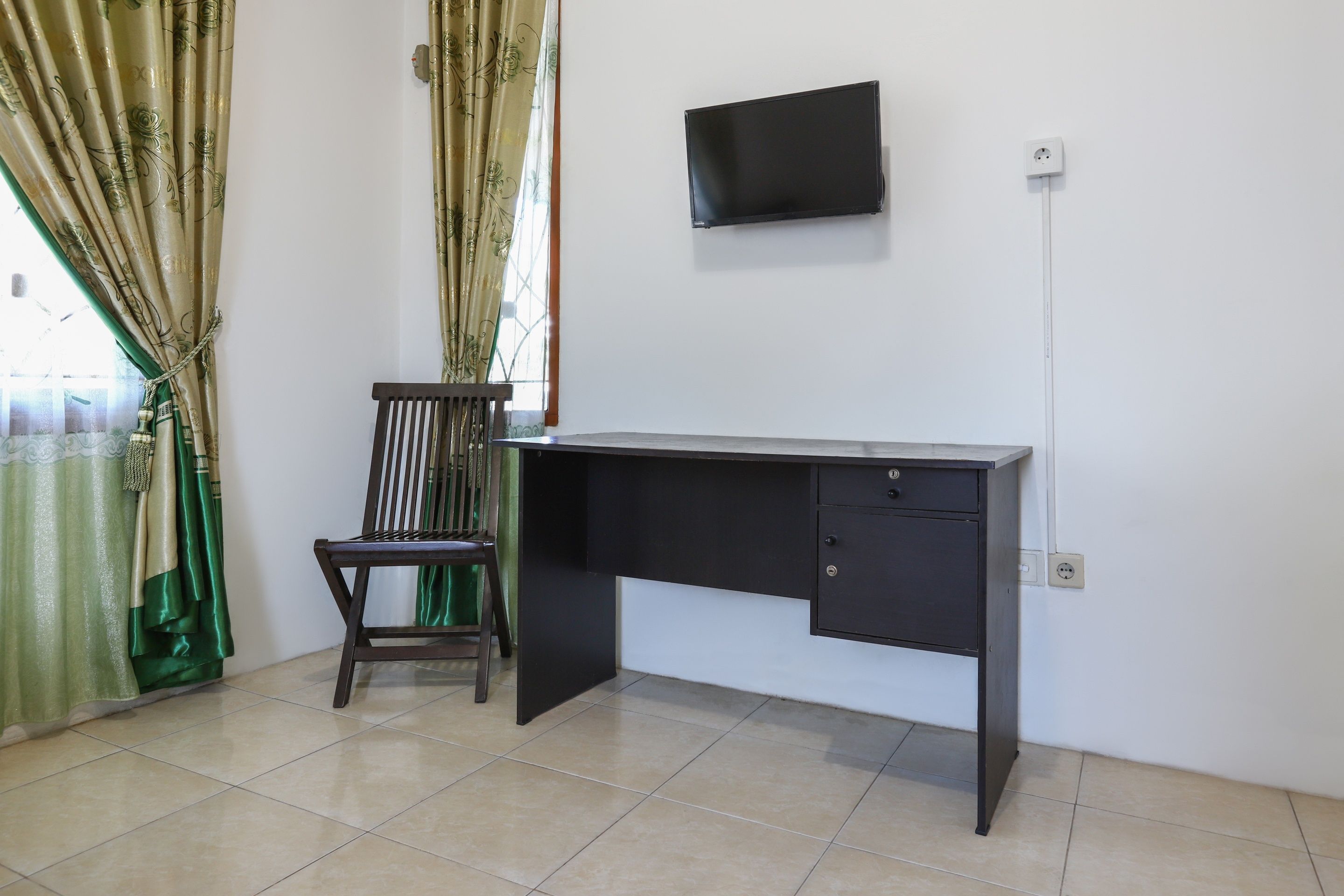 Una habitación con escritorio, silla, televisor montado en la pared y ventana con cortinas.