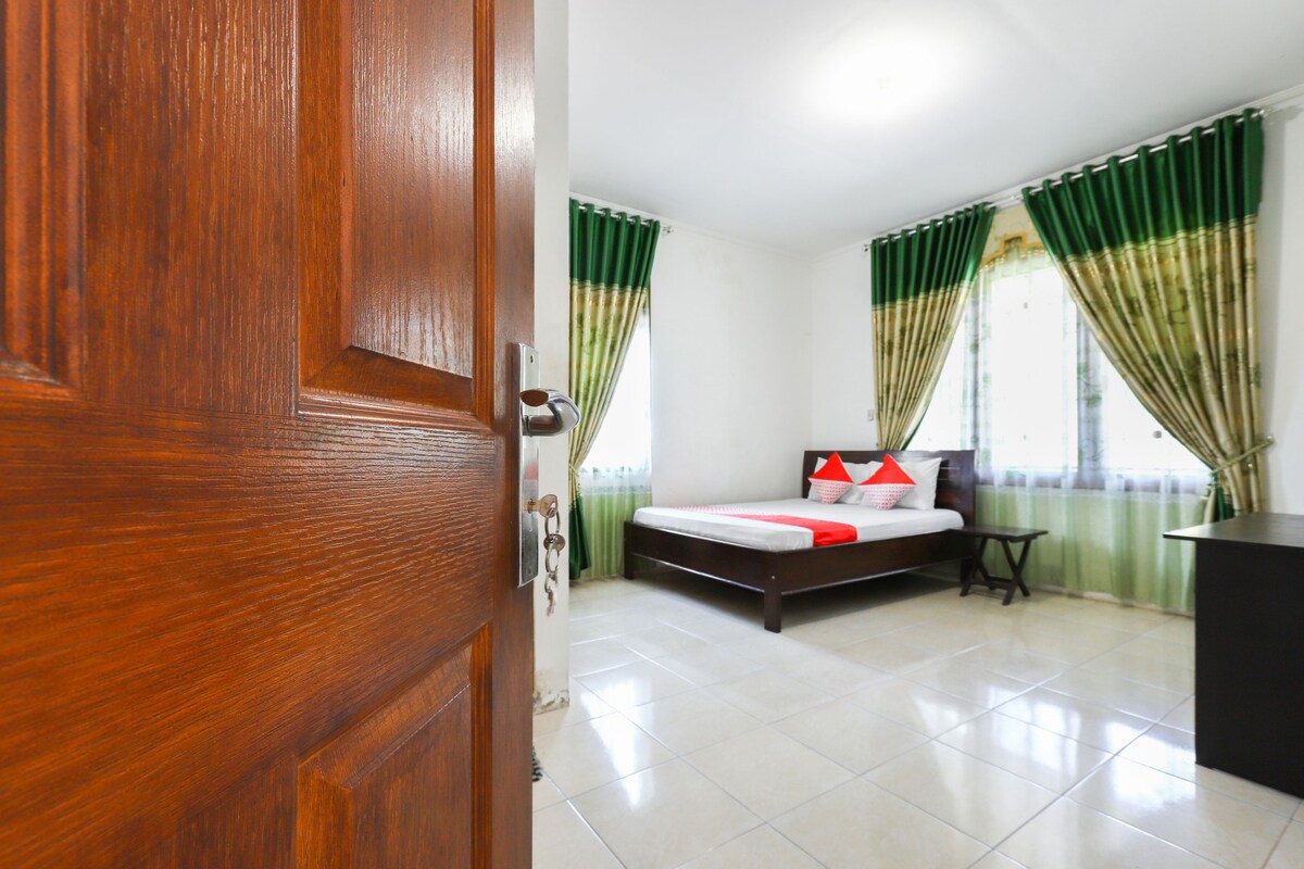Une chambre d’hôtel avec une porte en bois, un lit recouvert d’une courtepointe rouge et blanche, des rideaux verts et une petite table.