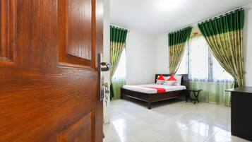 Una habitación de hotel con una puerta de madera, una cama con ropa blanca y almohadas rojas, cortinas verdes y piso de baldosas.