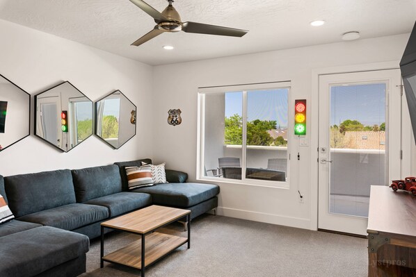 A modern living room with a sectional sofa, a coffee table, and a ceiling fan. There is a large window with a view of trees and a red traffic light outside.