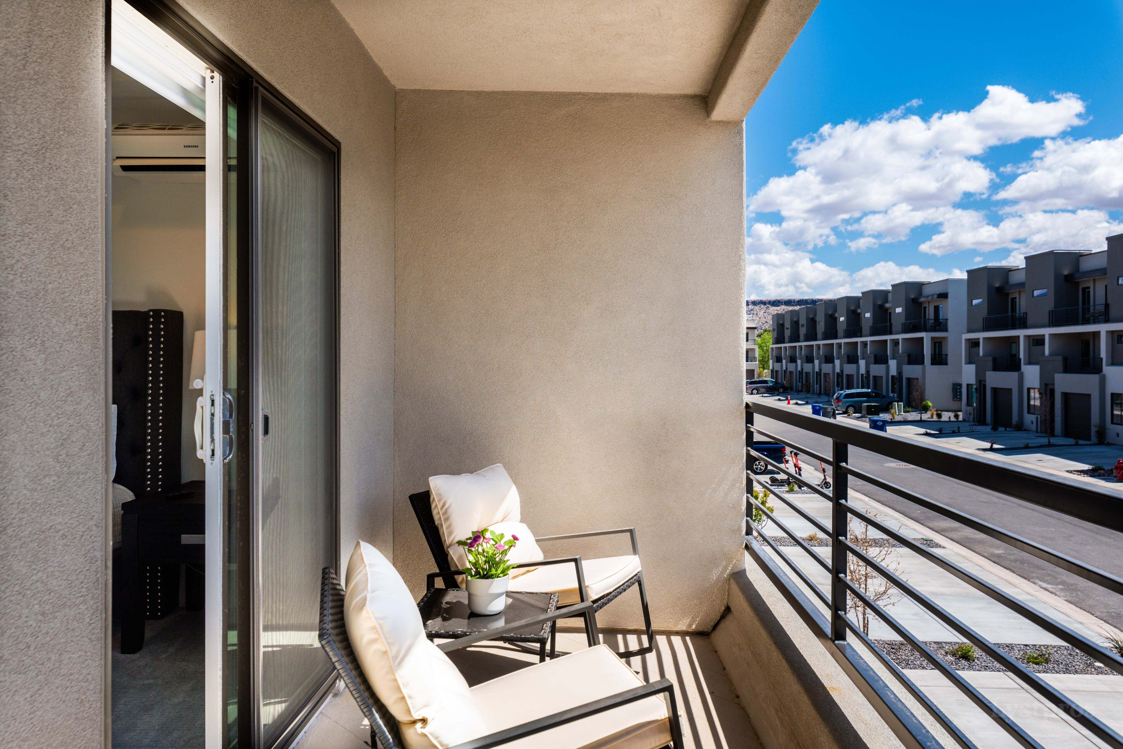 A balcony with two chairs and a small table with a plant, overlooking a street with parked cars and buildings.