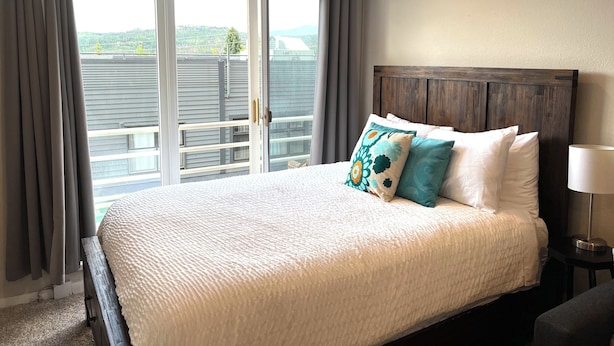 A hotel room with a large bed, a wooden headboard, and a view through a sliding glass door to the outside.