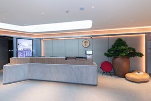 Un mostrador de recepción moderno con una pantalla digital, una planta grande en maceta y un taburete de madera original.