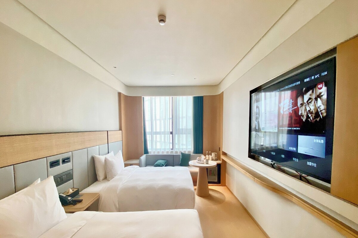 A hotel room with two beds, a TV on the wall, a small table, and a window with curtains.