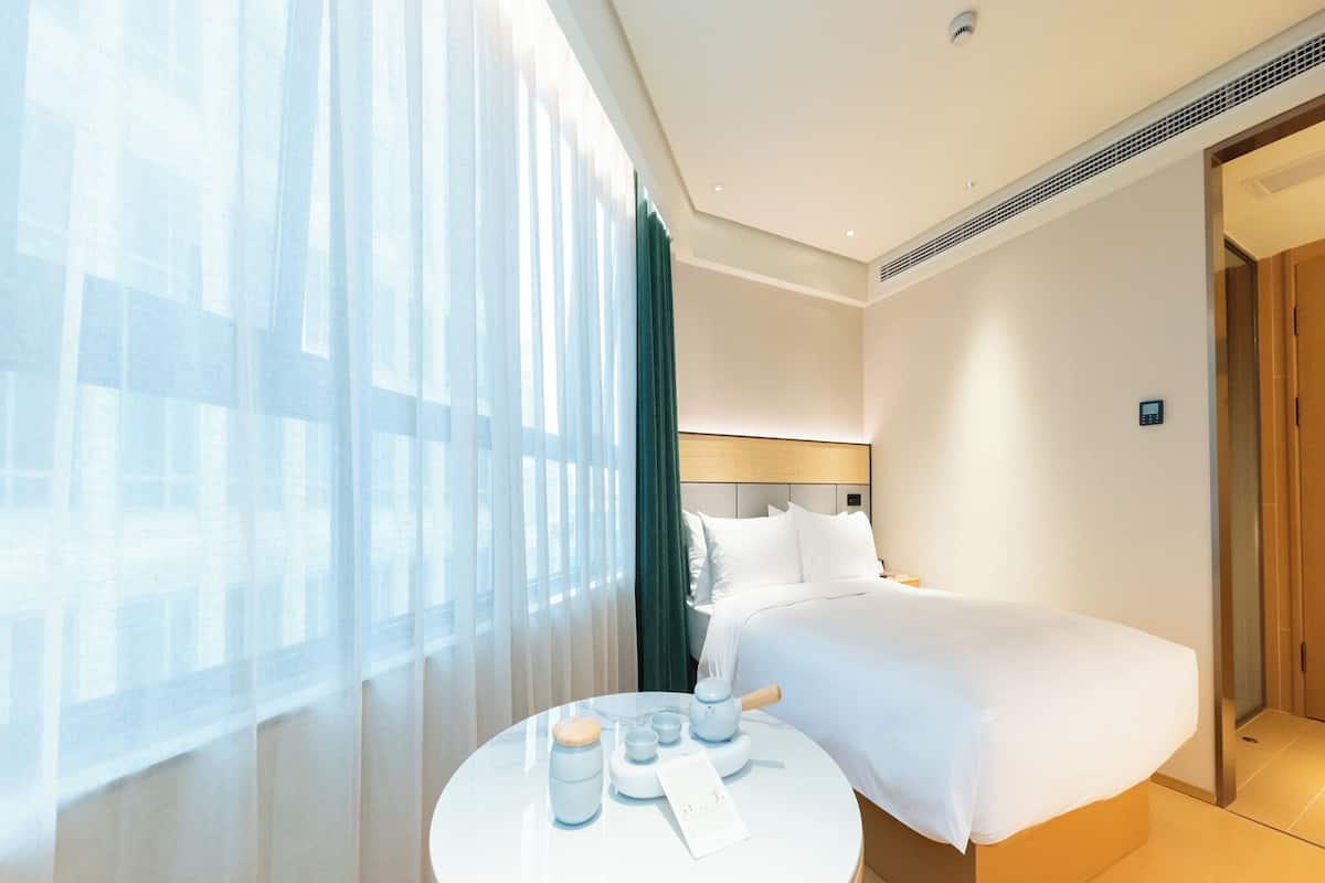 Habitación de hotel con cama, una mesa redonda con juego de té y vistas de la ciudad a través de cortinas translúcidas.