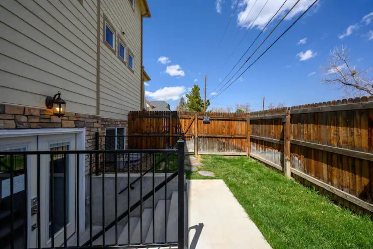 A residential backyard with a wooden fence, a paved pathway, and a house with siding and a stone foundation.