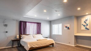 A bedroom with a bed, a nightstand, a window with blinds, a ceiling light, and a wall-mounted shelf with artwork.