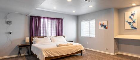 A bedroom with a bed, a nightstand, a window with blinds, a ceiling light, and a wall-mounted shelf with artwork.
