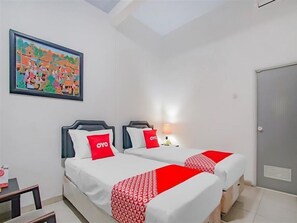 A hotel room with two beds, each with a red OYO branded pillow and blanket. There is a painting on the wall depicting a colorful scene, and a door on the right side of the room.