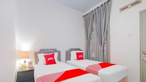 A hotel room with two beds, each with a red and white bedspread and a pillow with the 'OYO' logo. There is a nightstand with a lamp and a small table with a book and a card. The room has a window with grey curtains and a door with a sign on it.