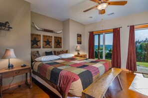 A bedroom with a large bed, a wooden bench, a ceiling fan, and a view of the outdoors through large windows.