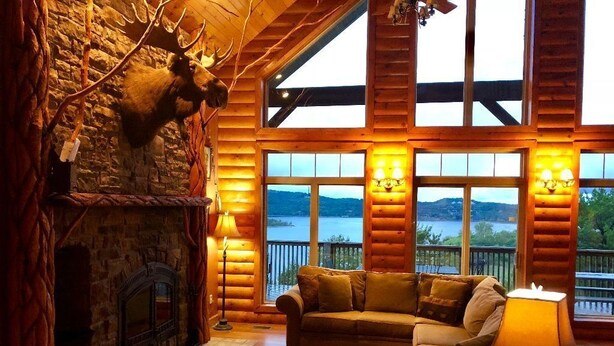 A log cabin interior with a stone fireplace, a green sofa, and a view of a lake through large windows. - Beautiful Views from the Rusty Moose (Branson West)