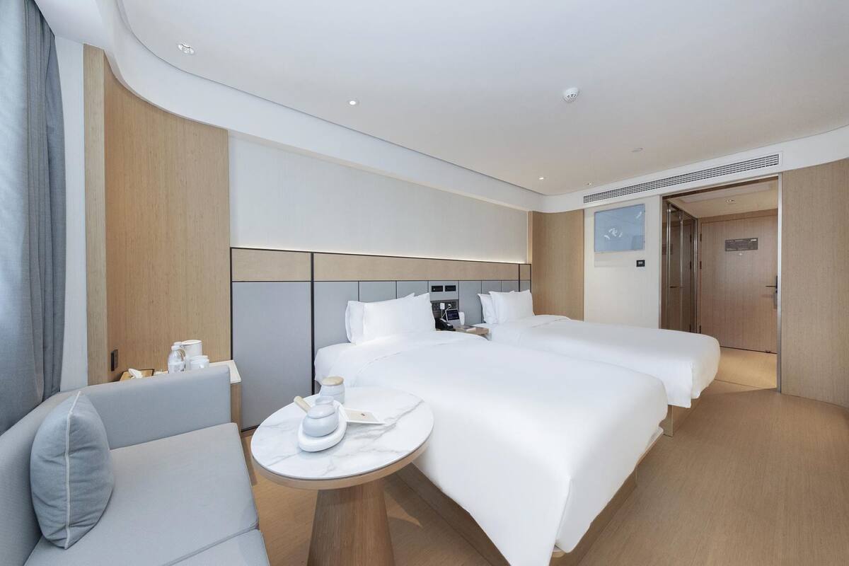 Habitación de hotel con dos camas, una mesita pequeña con hervidor, una silla y cabecera de madera.