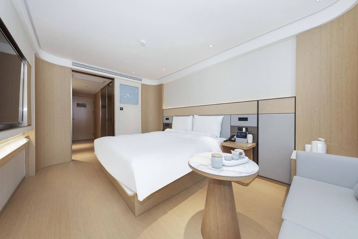 Una habitación de hotel moderna con una cama grande, una mesita de noche con lámpara, una pequeña mesa redonda con una taza y un platillo, y una silla.