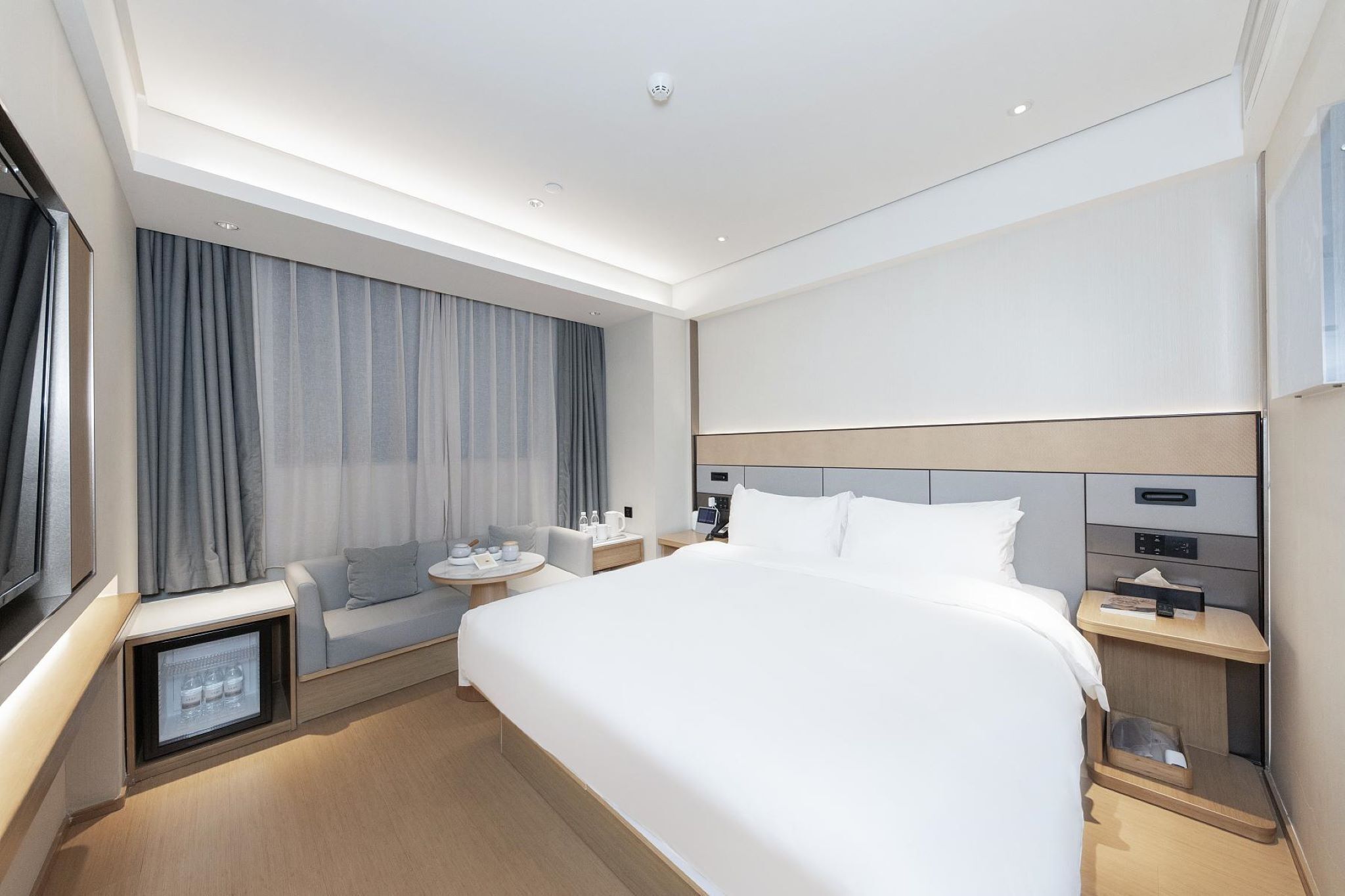 Moderni hotellihuone, jossa on suuri sänky, yöpöydät, pieni oleskelualue ja sisäänrakennettu minijääkaappi.