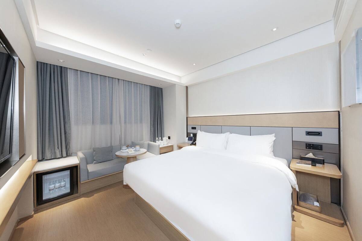 Una habitación de hotel moderna con una cama grande, mesitas de noche, una pequeña zona de estar y un mini refrigerador empotrado.