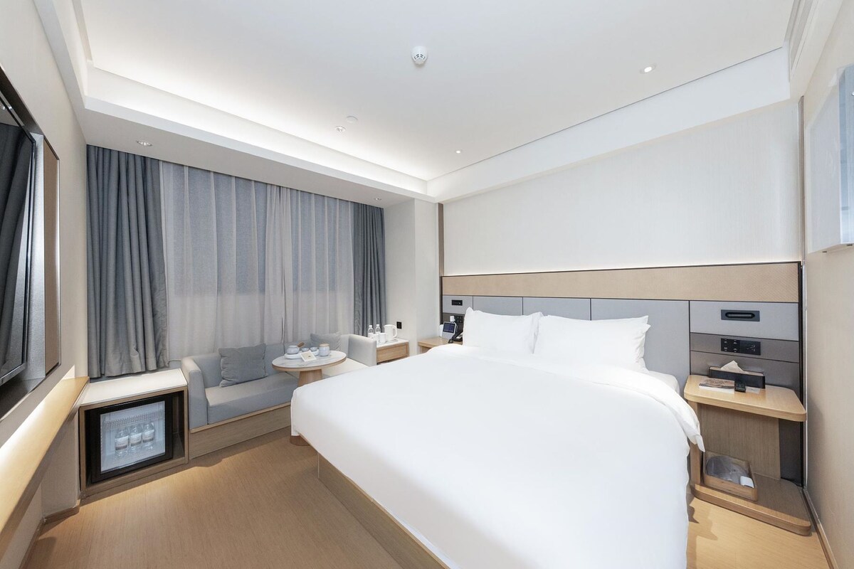 A modern hotel room with a large bed, bedside tables, a small seating area, and a built-in mini-fridge.