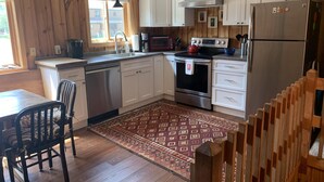 A kitchen with wooden walls, white cabinets, stainless steel appliances, a dining area with a chair, and a patterned rug.