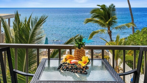 A glass table with fruits and a pineapple on a balcony overlooking the ocean. - OCEANFRONT; Stunning Corner Unit in Quiet Boutique Complex, Spacious, Pool, A/C! (Kailua-Kona)