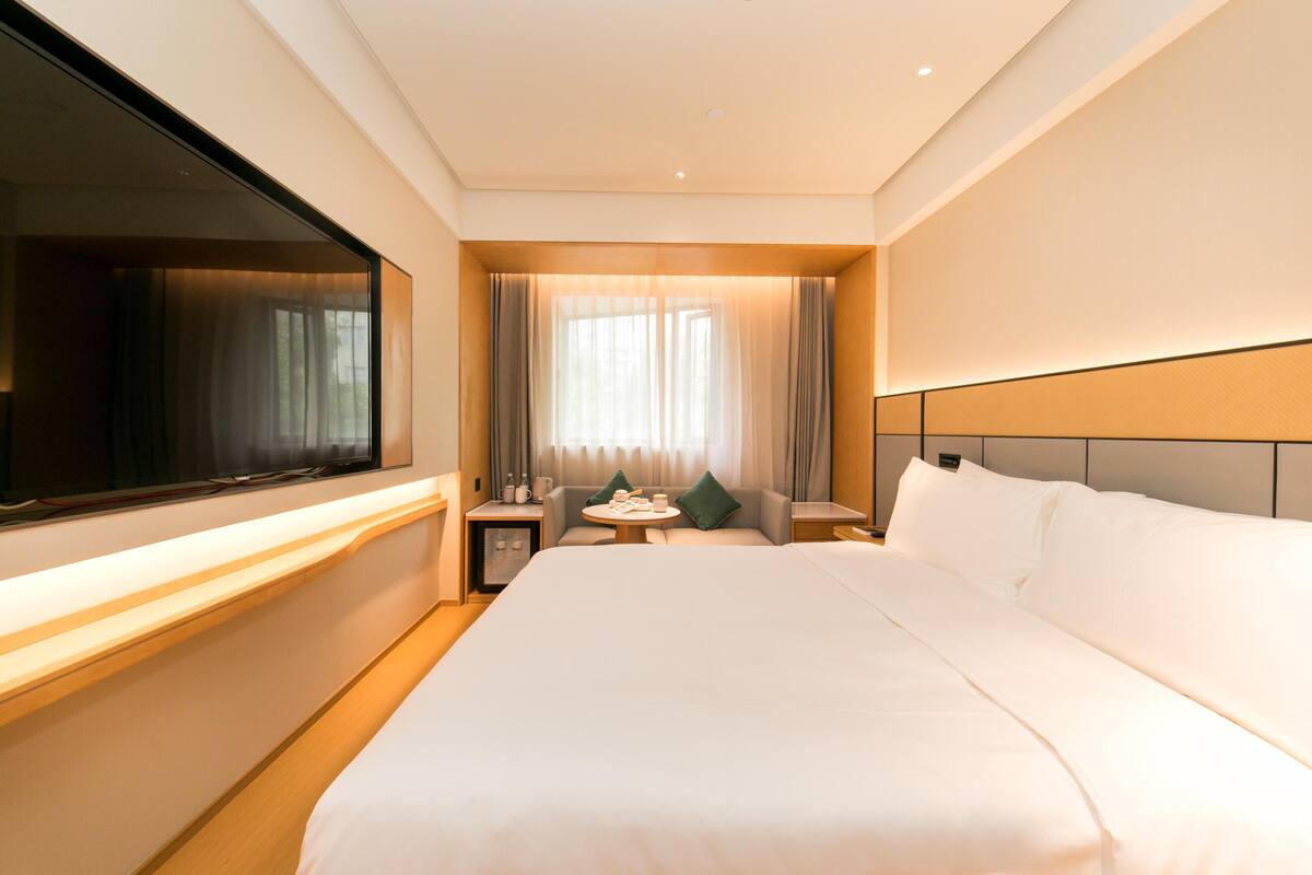 A hotel room with a large bed, a TV, a small table with a chair, and a window with curtains.