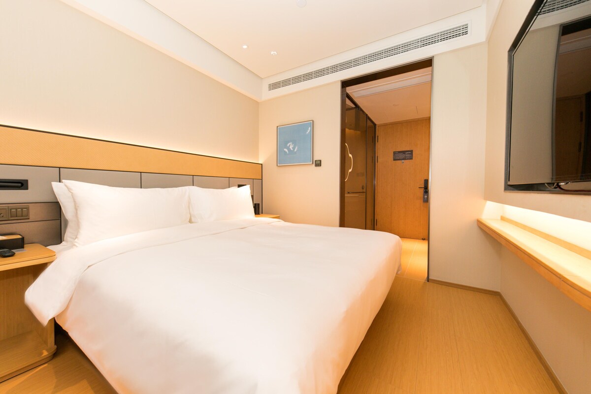 A hotel room with a large bed, a nightstand, a bedside table, a wall-mounted TV, and a door leading to another room.