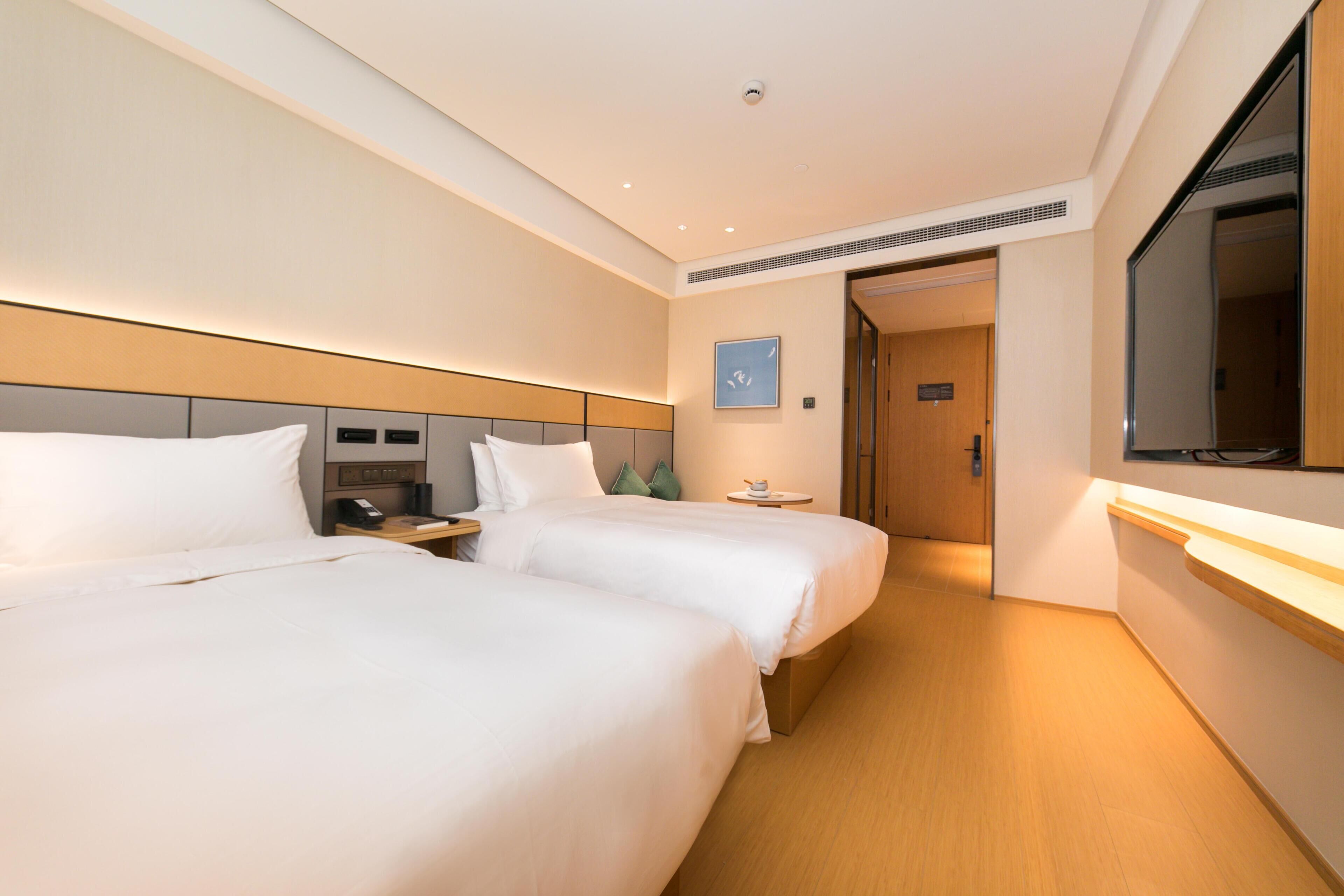 Habitación de hotel con dos camas, mesitas de noche, una pequeña mesa con una taza, un televisor montado en la pared y una puerta que da a otra habitación.
