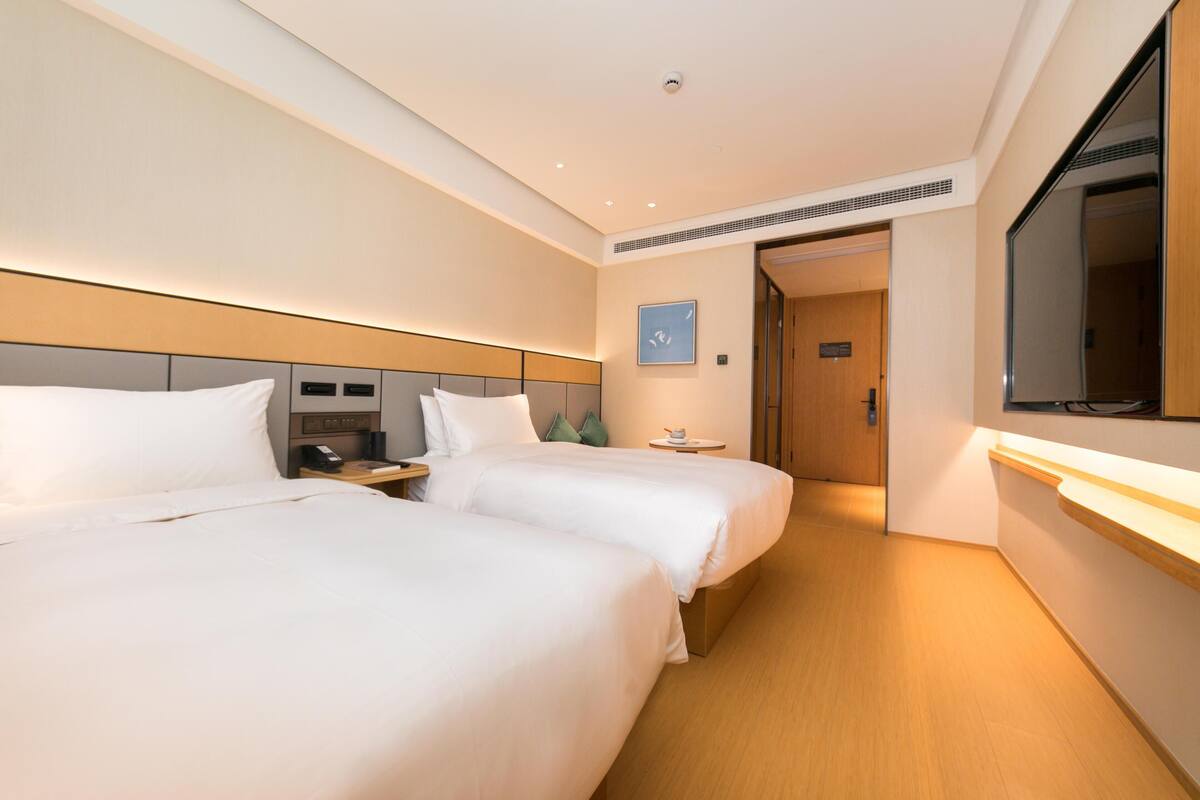 A hotel room with two beds, bedside tables, a small table with a cup, a wall-mounted TV, and a door leading to another room.