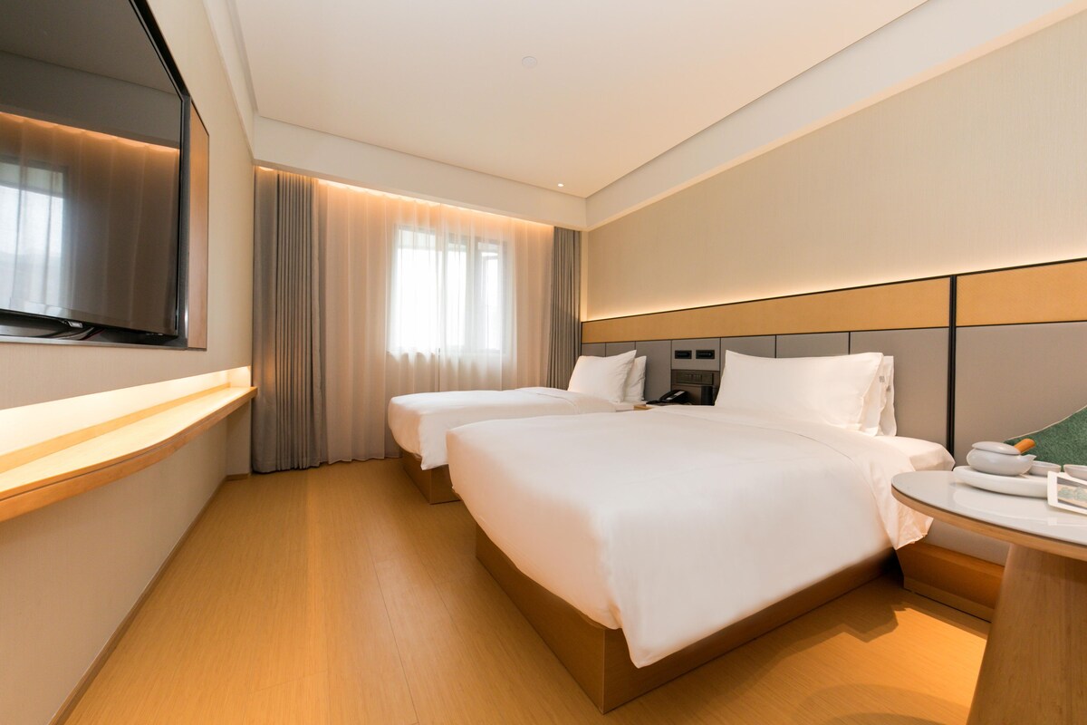 A hotel room with two beds, a flat-screen TV, a bedside table, and a window with curtains.