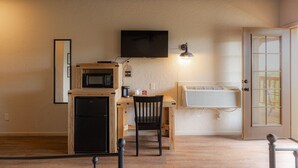 Een functionele keuken met een magnetron, koelkast en een eettafeltje met een stoel. Een flatscreen televisie is aan de muur boven de tafel bevestigd, en er is een raamairconditioner in de muur geplaatst.
