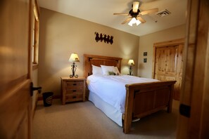 A bedroom with a wooden bed, two bedside tables with lamps, a ceiling fan, and a wooden door.