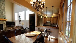 Un comedor espacioso con una mesa y sillas de madera, una chimenea de piedra y una cocina con armarios de madera e iluminación colgante.