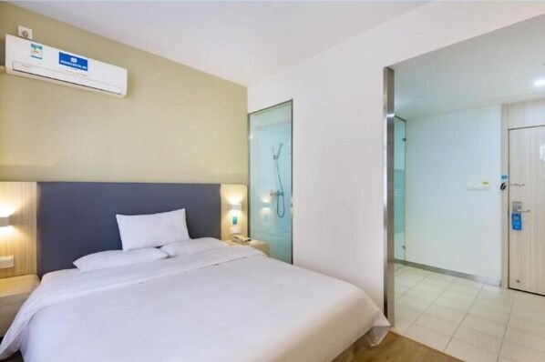 A hotel room with a bed, bedside lamp, nightstand, and a wall-mounted air conditioner.