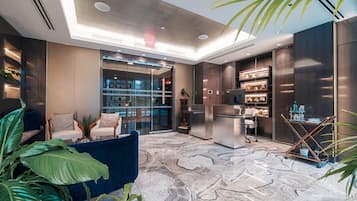 A modern hotel lobby with a marble floor, seating area, and a kitchen visible through glass doors.