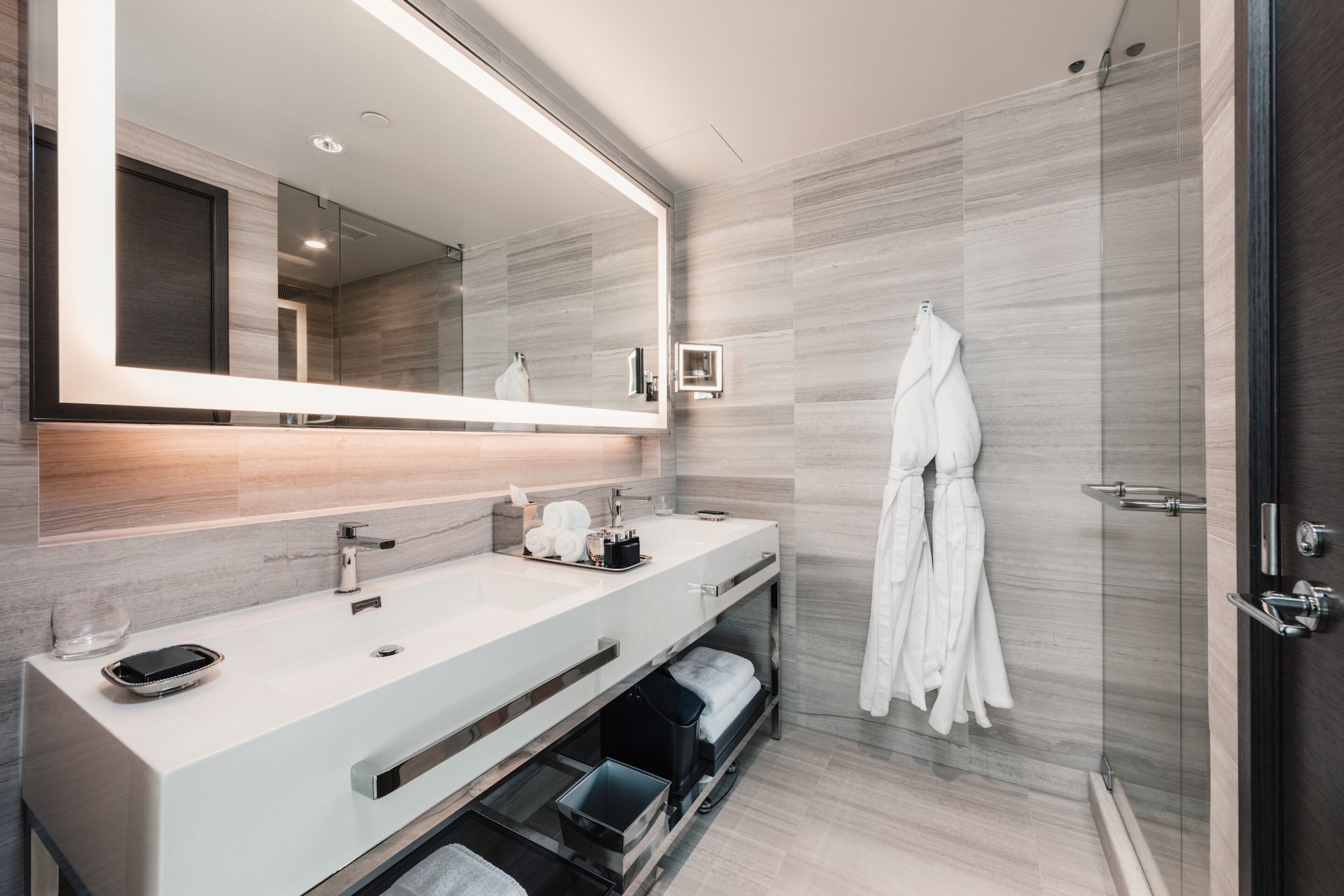 A modern bathroom with a large mirror, two sinks, a glass shower enclosure, and a robe hanging on the wall.