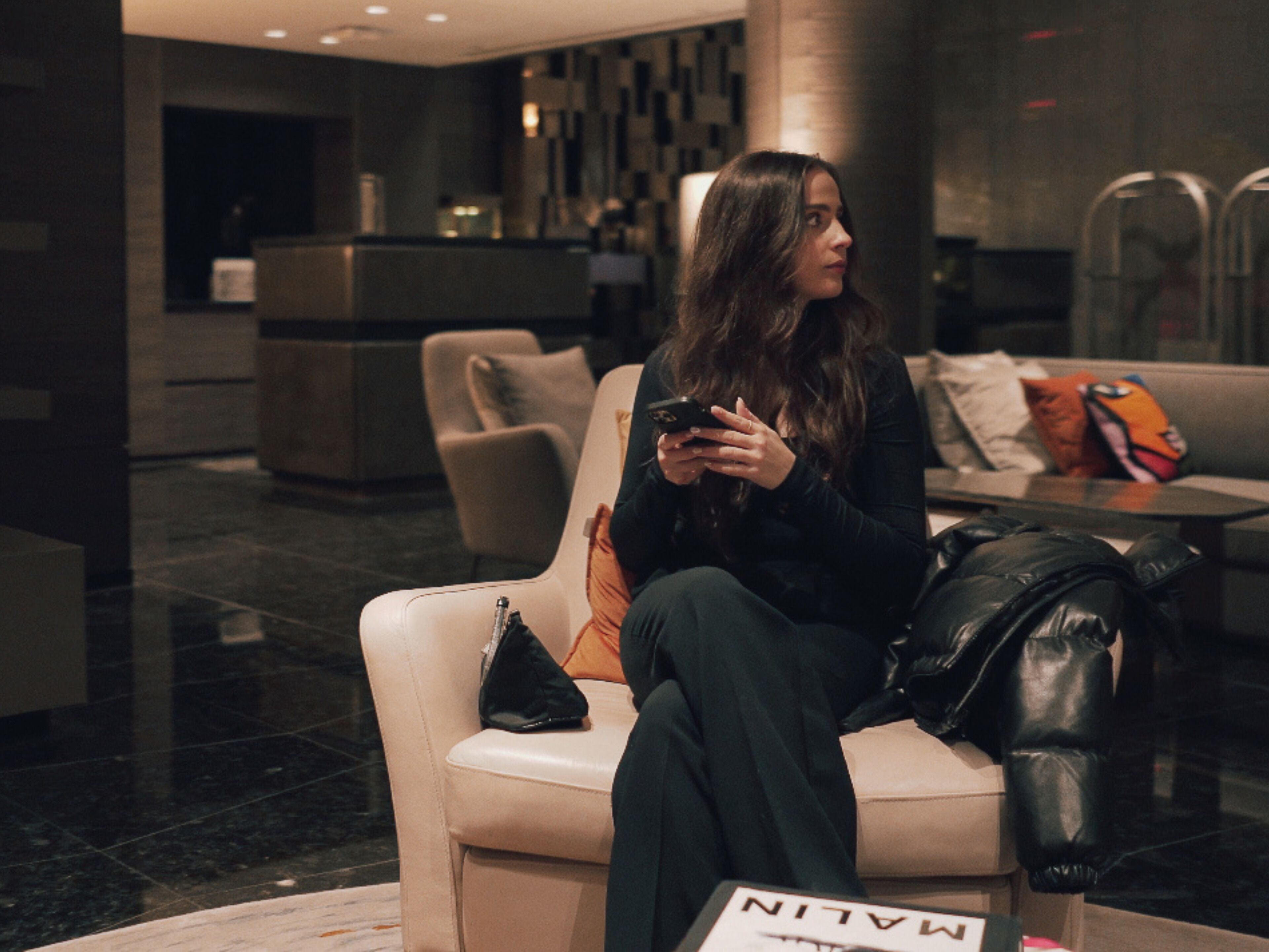 A woman sitting on a leather couch in a lobby with a black handbag beside her.