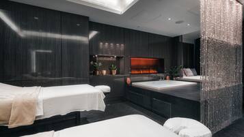 A modern spa room with two beds, a large mirror, a fireplace, and a decorative wall.