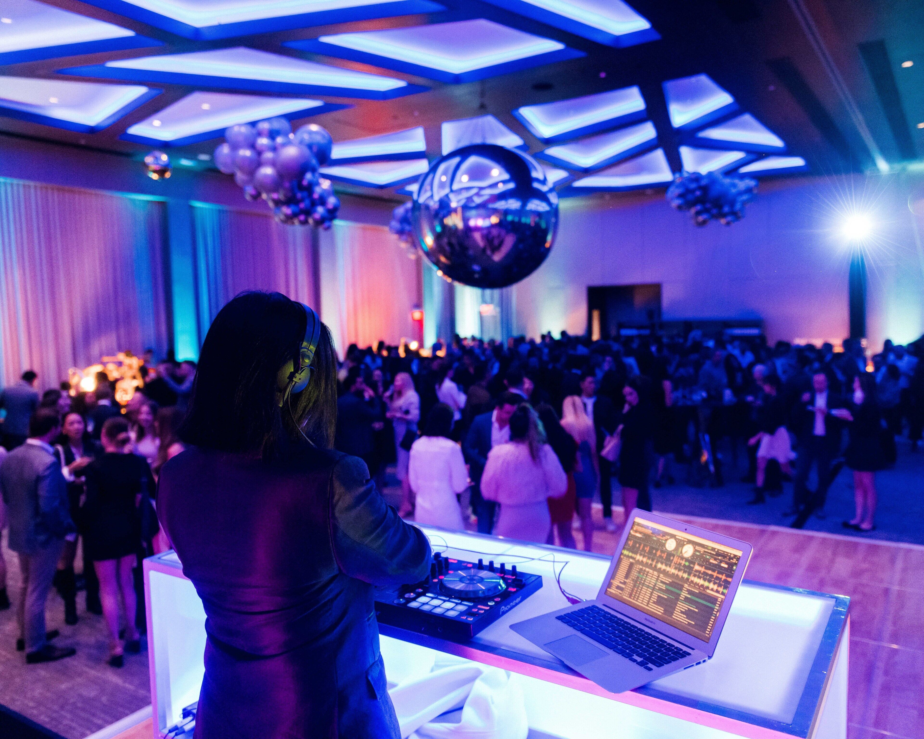 Uma pessoa em uma cabine de DJ com um laptop e equipamentos de DJ, cercada por uma multidão em um salão com design de teto moderno.