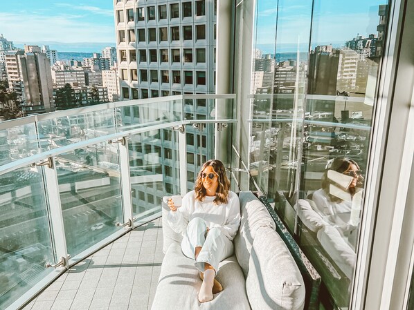 A person sitting on a white couch with a glass railing, overlooking a cityscape with tall buildings.