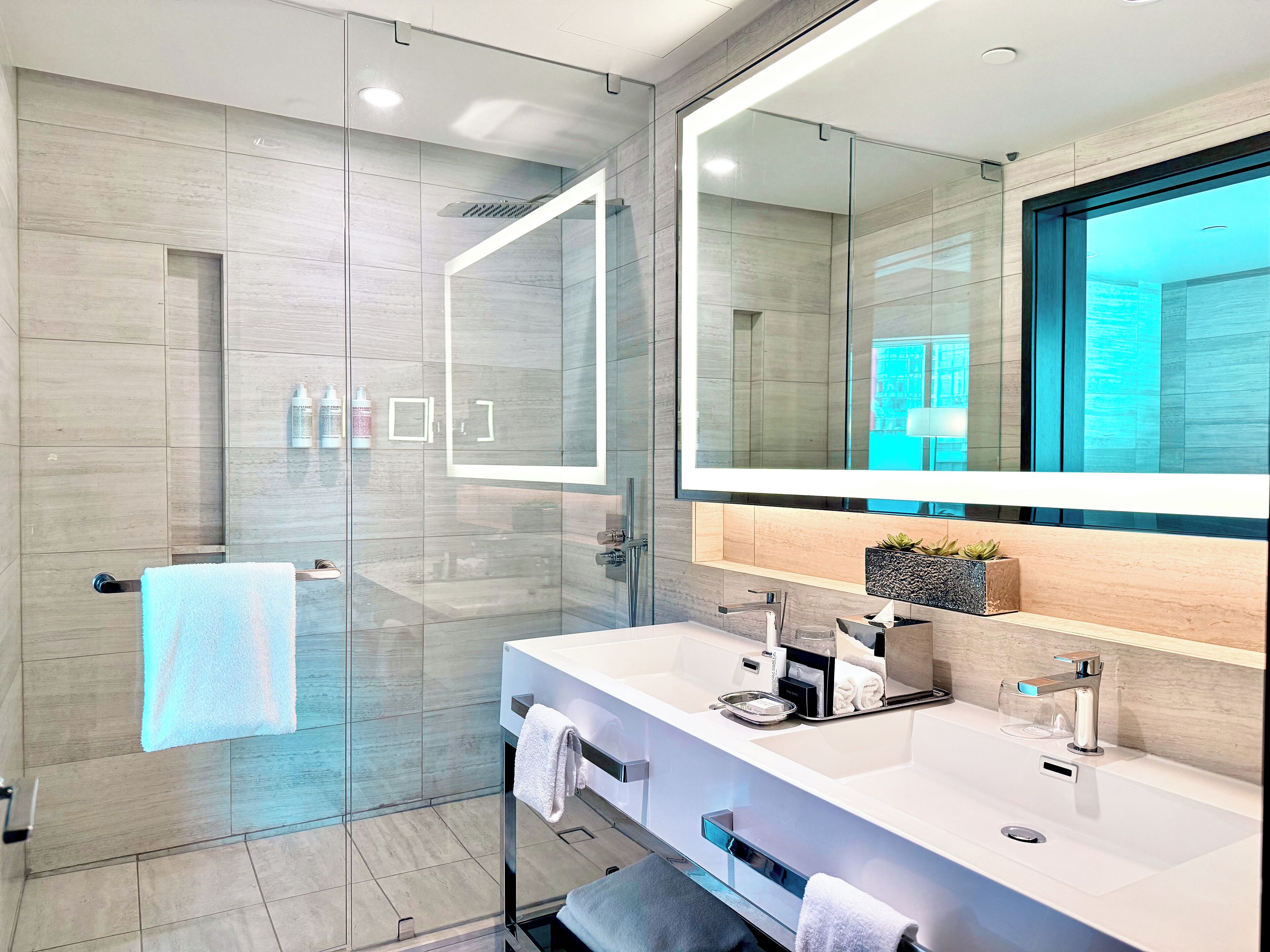 A modern bathroom with a glass shower enclosure, a large mirror, and a vanity with dual sinks and a towel rack.