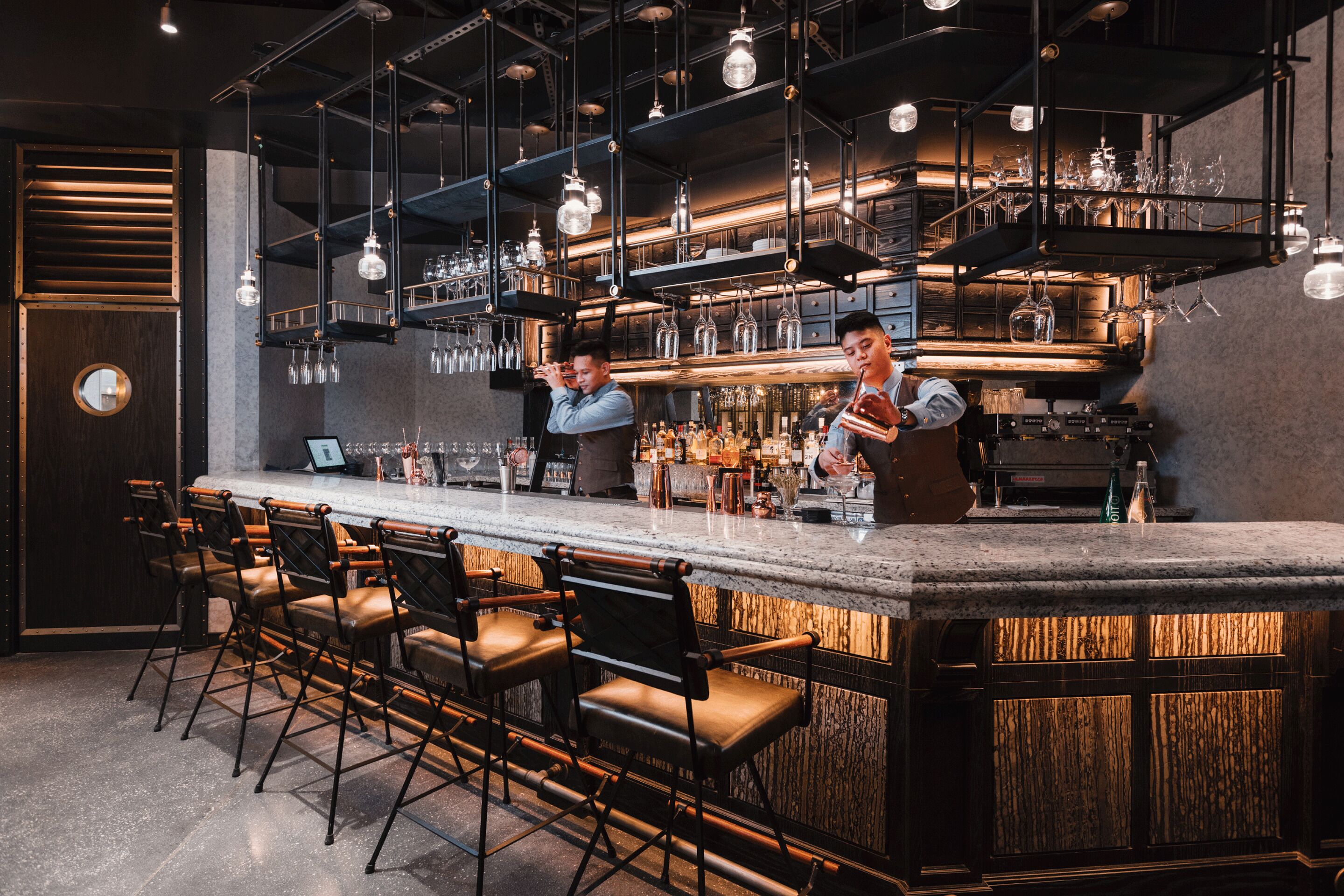 A bar area with a long counter, multiple stools, and hanging light fixtures.