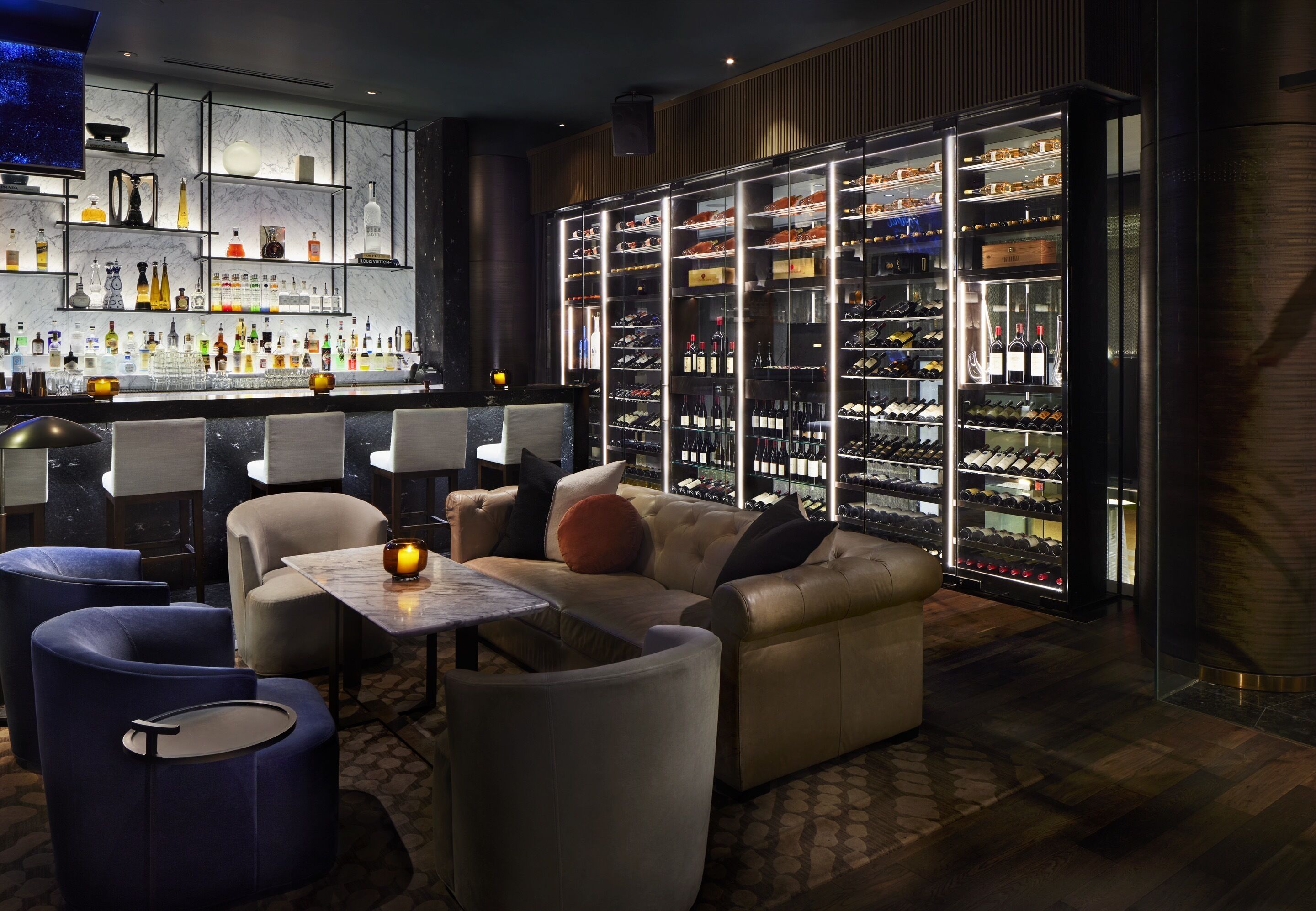 A bar area with a marble countertop, shelves with bottles, and a wine cooler. There are sofas and a table with a candle.
