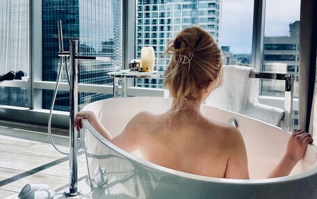 A woman relaxing in a bathtub with a city view, a towel, and a small table with items.
